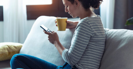 woman sat on sofa looking at phone in one hand and holding a cup of tea in another