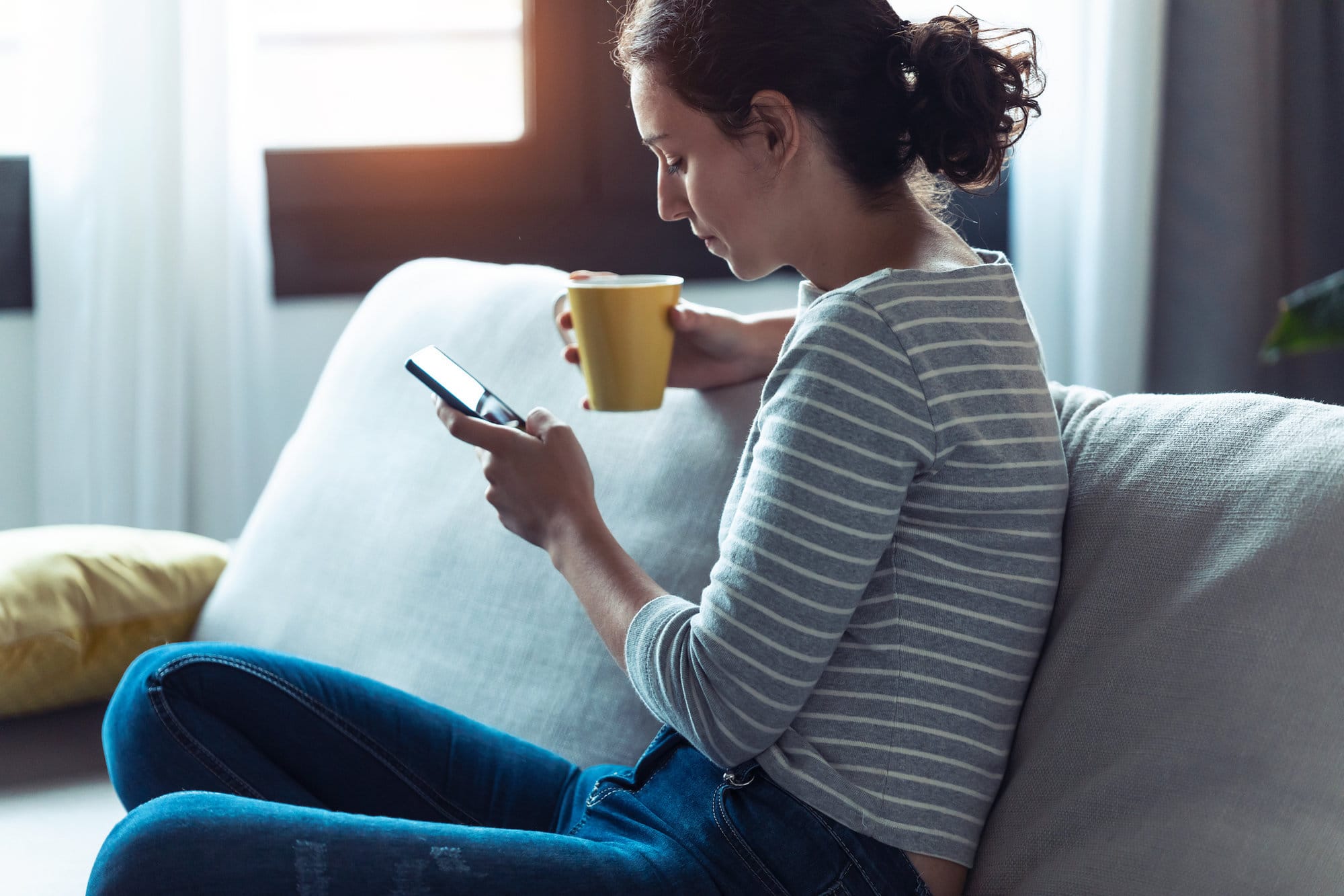 woman sat on sofa looking at phone in one hand and holding a cup of tea in another