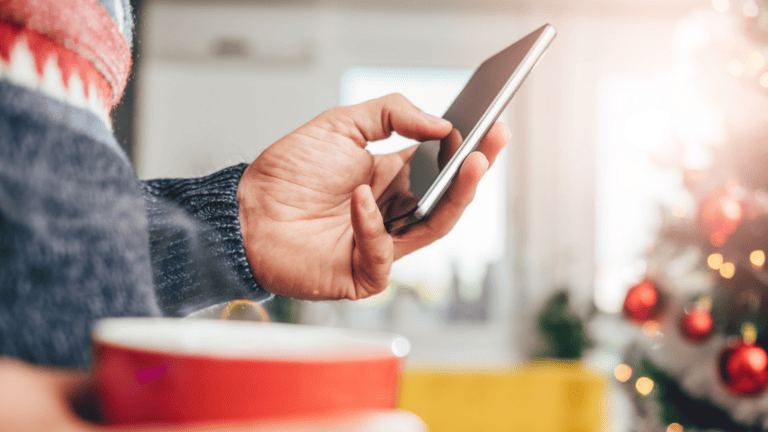 A man wearing a Christmas jumper scrolling through his phone and holding a cup of tea with a Christmas tree in the background