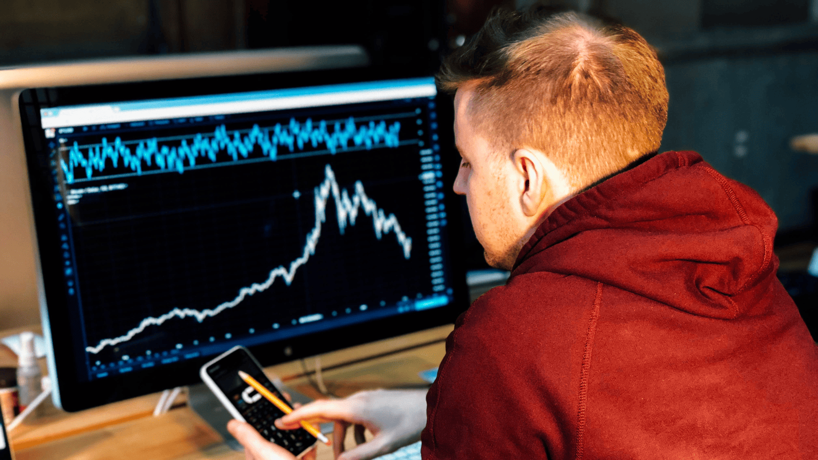 A man looking at his phone with his desktop showing crypto graphs in front of him