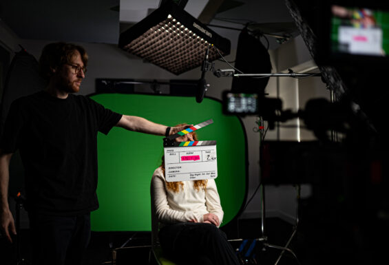 A young person in front of a green screen with a film crew