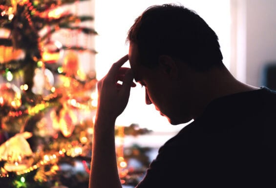 A man with his hand on his head looking stressed in front of a lit Christmas tree.