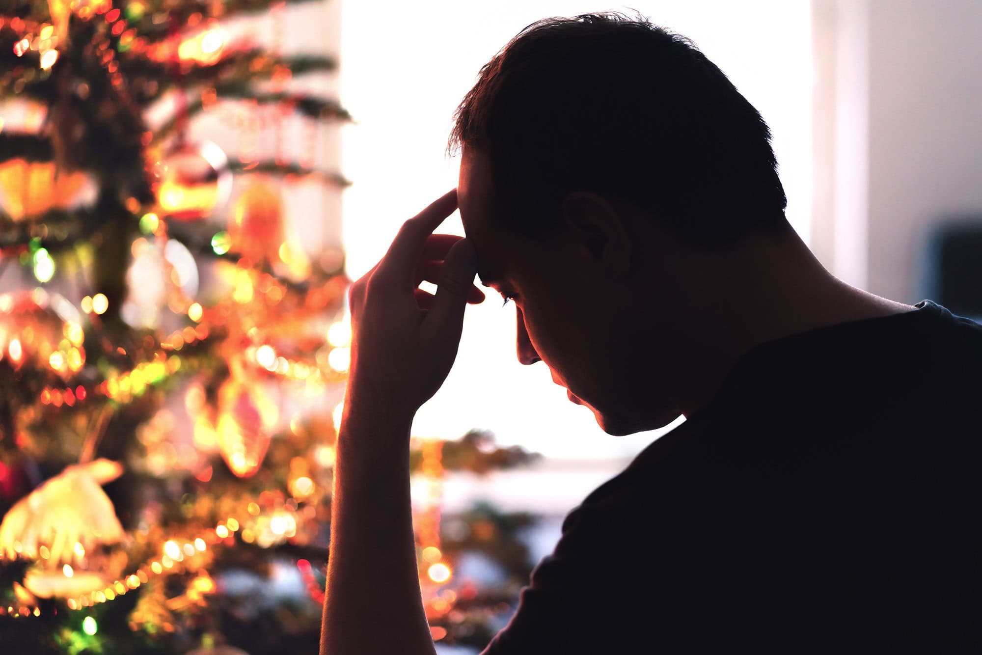 A man with his hand on his head looking stressed in front of a lit Christmas tree.